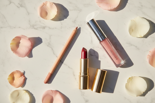 A premium beauty flat lay featuring a nude lip liner pencil, pink lip gloss, and berry lipstick arranged on white marble with rose petals — ideal for a lip makeup and lip care tutorial.